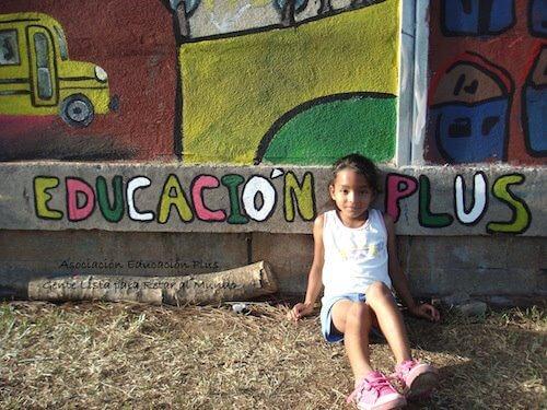 Niña sentada contra una pared que tiene pintado el texto Educacion Plus.