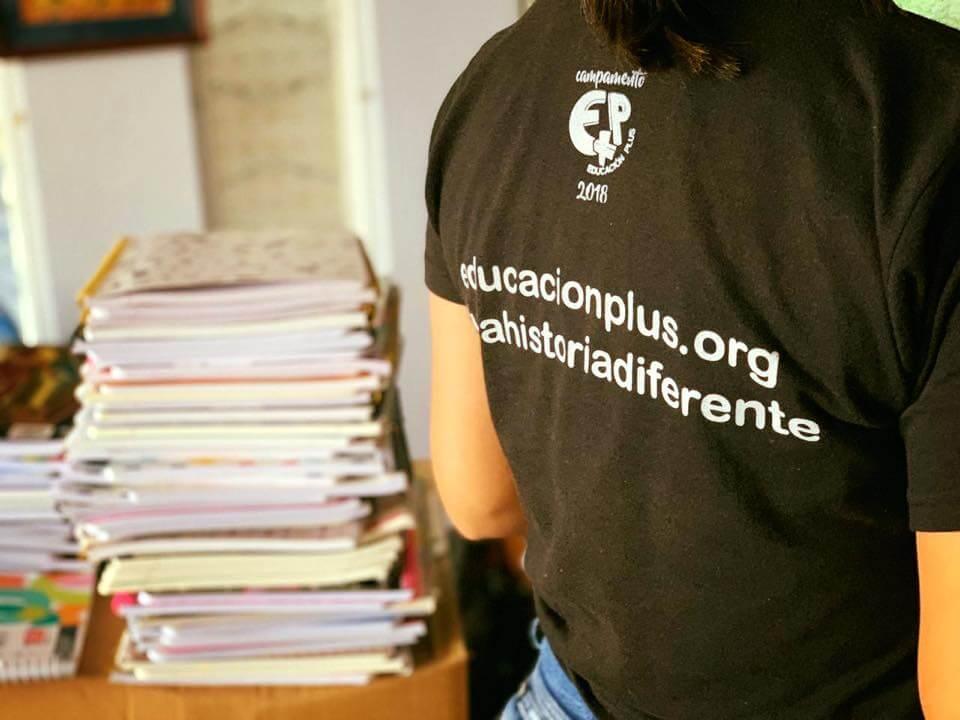 Cuadernos apilados sobre una mesa u una persona de espaldas con una camisa de Educacion Plus.